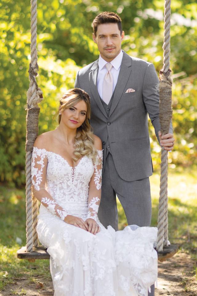 groom wearing Michael Kors Medium Grey Performance suit standing behind the bride in a lace dress sitting on a large outdoor swing with greenery in the background.